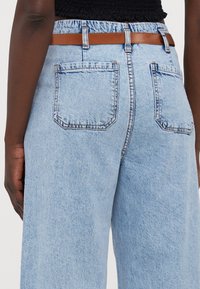 Jeans de ganga de cintura alta azul claro com um cinto de couro castanho, apresentando dois bolsos traseiros e uma textura levemente desbotada.