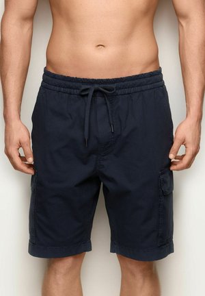 Mężczyzna ubrany w granatowe szorty cargo z obwiązywaną talią, stojący na tle jednolitego jasnego tła.