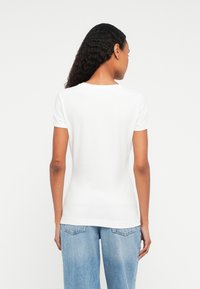 Frau mit langen lockigen Haaren, die ein schlichtes weißes T-Shirt mit kurzen Ärmeln und weit geschnittene hellblaue Jeans trägt, steht mit dem Rücken zur Kamera.