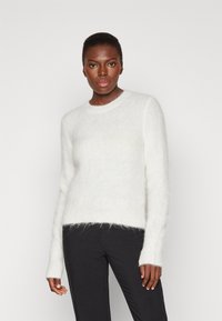 Maglione bianco peloso con scollatura rotonda, maniche lunghe e una consistenza morbida. Indossato con pantaloni neri, caratterizzati da dettagli di design minimi.
