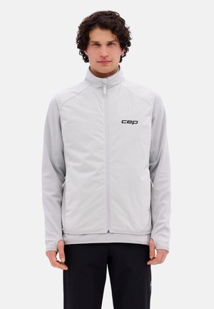 Man wearing light grey zip-up vest with "cep" logo over long-sleeve shirt and black pants, standing against white background.