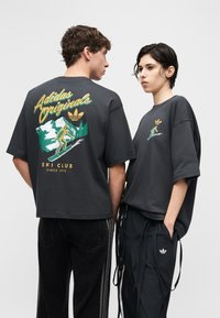 To personer iført mørke Adidas Originals ski klubb T-skjorter med skigrafikk, stående side om side mot en ensfarget bakgrunn.