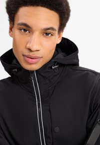 Zwarte hoodie met een rits aan de voorkant, gemaakt van een gestructureerde stof, met witte piping langs de rits en verstelbare toggles aan de capuchon.