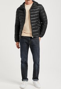 Man i en svart puffjacka över en beige tröja, blå jeans och vita sneakers, stående mot en ljus, enkel bakgrund.