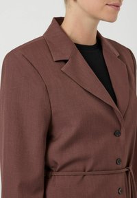 Vrouw in een bruine blazer met een riem over een zwart ribbeltop, van schouders tot midden torso op een effen achtergrond.