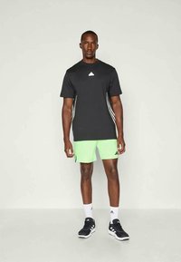 Camisa negra de manga corta con logo blanco, combinada con pantalones cortos verdes neón que presentan cortes laterales distintivos, llevada con zapatillas deportivas negras y calcetines blancos.