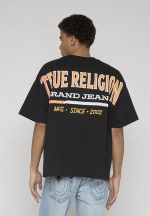 Mężczyzna z kręconymi włosami w czarnej, oversize'owej koszulce z napisem „True Religion Brand Jean MFG Since 2002” na plecach oraz w jasnoniebieskich dżinsach.