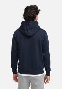 Un hoodie bleu marine en mélange de coton doux, avec une grande capuche ajustable et des poignets et un ourlet côtelés, porté par-dessus une chemise blanche.