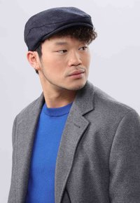 Dark blue flat cap, wool-blend, with stitching detail. Gray textured blazer over a blue sweater, creating a layered look.