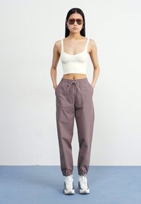 Žena stoji samouvjereno protiv jednostavne pozadine, odjevena u bijeli crop top i sivo donje trenerke, uz dodatke poput sunčanih naočala i sportskih tenisica.