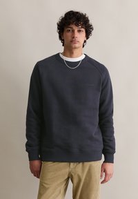 Felpa grigia scura con scollo a giro e polsini a costine, con un logo sottile sul petto. Indossata con pantaloni in corduroy beige chiaro.