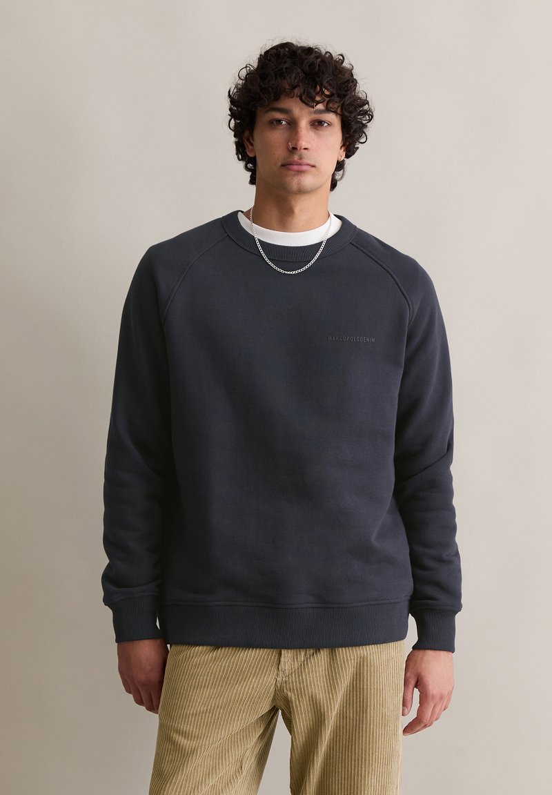 Felpa grigia scura con scollo a giro e polsini a costine, con un logo sottile sul petto. Indossata con pantaloni in corduroy beige chiaro.