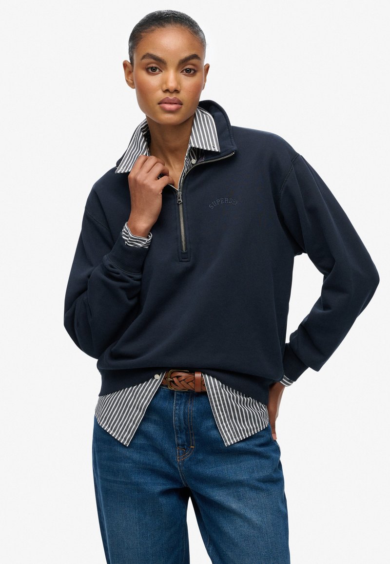 Marine kwart-rits sweatshirt met geribde manchetten en een logo, gelaagd over een wit en marine gestreept shirt, gecombineerd met blauwe jeans en een leren bruine riem.