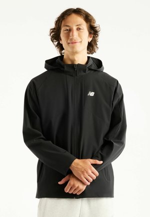 Jong persoon met golvend haar draagt een zwarte New Balance zip-up hoodie, handen voor zich geklemd, tegen een witte achtergrond.