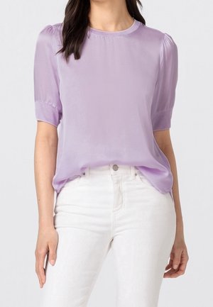 Blouse lavande en tissu léger avec manches courtes bouffantes. Elle présente un col rond et une coupe décontractée, associée à un jean blanc.