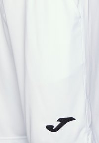 Pantalons de sport blancs en tissu lisse et léger avec un logo noir brodé sur le côté et des coutures discrètes.