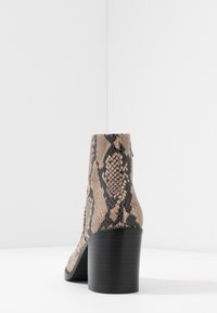 Snakeskin-mönstrad ankelboot i beige och svart, med en texturerad yta och en kraftig svart klack, visad bakifrån mot en vit bakgrund.