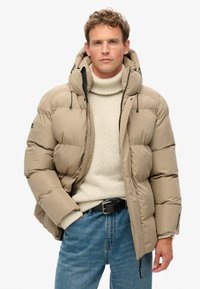 Giacca puffer beige con cappuccio, dotata di tasche con zip e cordini regolabili. Indossata sopra un dolcevita crema e jeans blu.