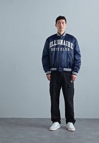 Muž stojí proti sivej stene, oblečený v tmavomodrom saténovom saku s nápisom "Billionaire Boys Club", čiernych cargo nohaviciach a bielych teniskách.