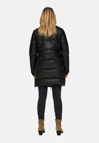 Zwarte pufferjack met een glanzende afwerking, knielang, horizontaal gewatteerd design, gecombineerd met donkere jeans en bruine enkellaarsjes met een hak.