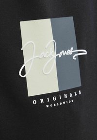 Crna tkanina središnjeg uzorka sa dva obojena kvadrata, svijetlozelene i plave boje, te bijelim izvezenim natpisom "Jack Jones Originals Worldwide."