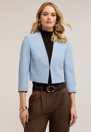 Vrouw met blond haar die oorbellen van goudblad draagt, lichtblauw cropped blazer, zwart topje, bruine hoge taille broek en gouden armband.