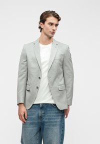 Γκρι σακάκι με υφή, δύο μπροστινά κουμπιά, καφέ πέτο και πλαϊνές τσέπες, φορεμένο πάνω από ένα λευκό πουκάμισο.