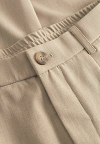 Közelkép bézs színű vászonnadrágról, amely gombos derékrésszel, rugalmas hátrésszel, övbújtatóval és részletes varrással rendelkezik.