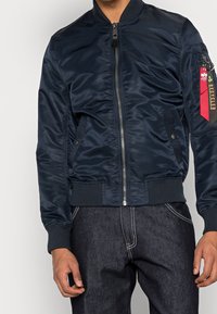 Giacca bomber blu navy in materiale liscio e lucido. Caratterizzata da collo e polsini a coste, chiusura frontale con zip e tasca laterale decorativa con etichetta.