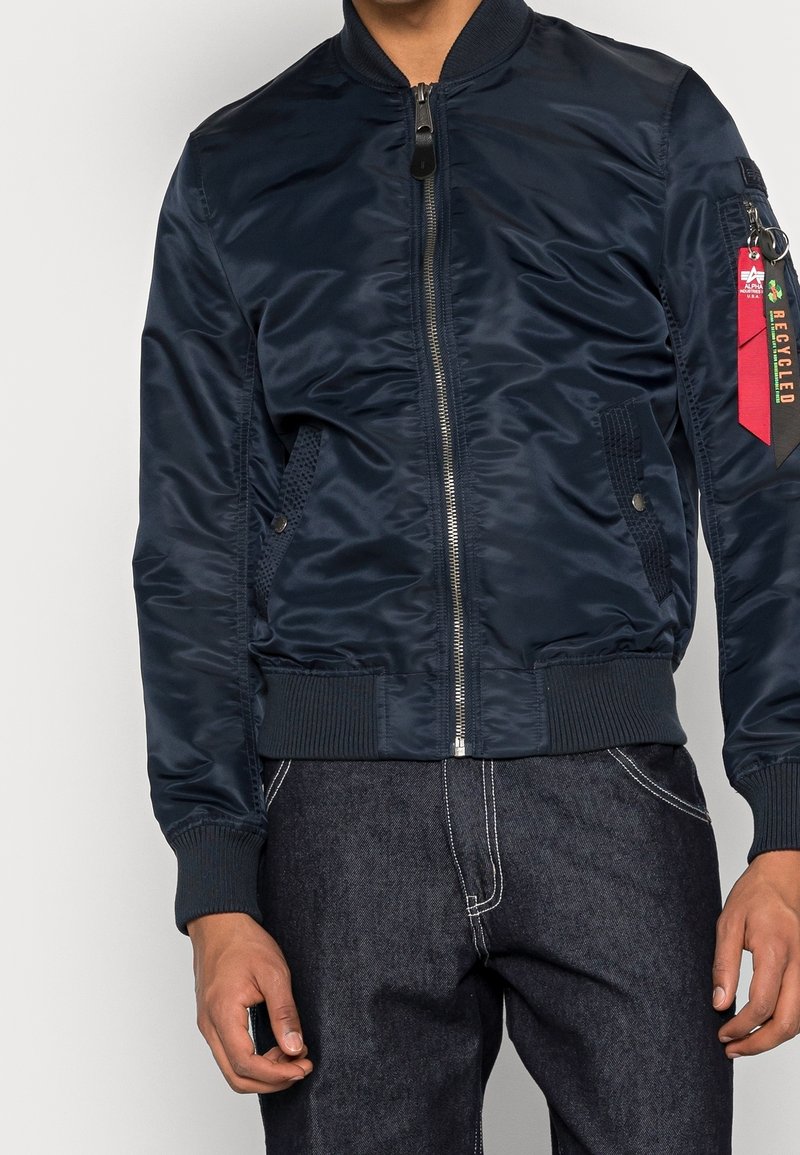 Giacca bomber blu navy in materiale liscio e lucido. Caratterizzata da collo e polsini a coste, chiusura frontale con zip e tasca laterale decorativa con etichetta.