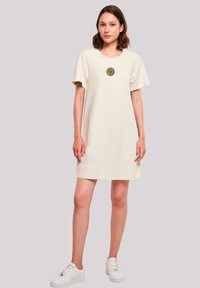 Crèmekleurig jurk met korte mouwen en een ronde halslijn, met een margrietafbeelding in het midden. G gedragen met witte sneakers. Gladde katoenen stof, tot de knie.