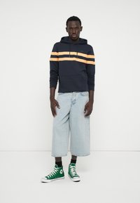 Navy hoodie with orange stripes, paired with light blue wide-leg jeans and green high-top sneakers. Simple design with pocket detail.