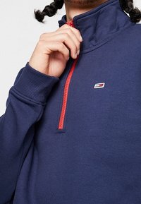 Mörkblå pullover-tröja med hög krage, röd dragkedja och liten logodetalj på bröstet. Mjuk bomullsblandning.