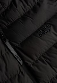 Nærbillede af sort quiltet stof med "SUPERDRY®" broderet og en sort lynlås-trækstang med "SUPERDRY" tekst.