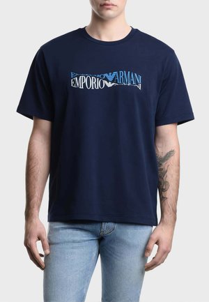 Uomo con T-shirt blu navy Emporio Armani con logo bianco e blu, abbinata a jeans azzurri, con tatuaggio sull'avambraccio sinistro.
