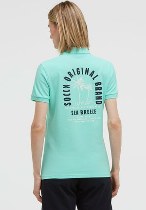Blonde Person trägt ein türkisfarbenes Polo-Shirt mit einer Palmen-Grafik und dem Text "SOCCX ORIGINAL BRAND SEA BREEZE" auf dem Rücken.