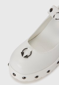 White leather shoe with a rounded toe, adjustable strap, silver circle hardware, and pointed accents; features a thick platform with studs.