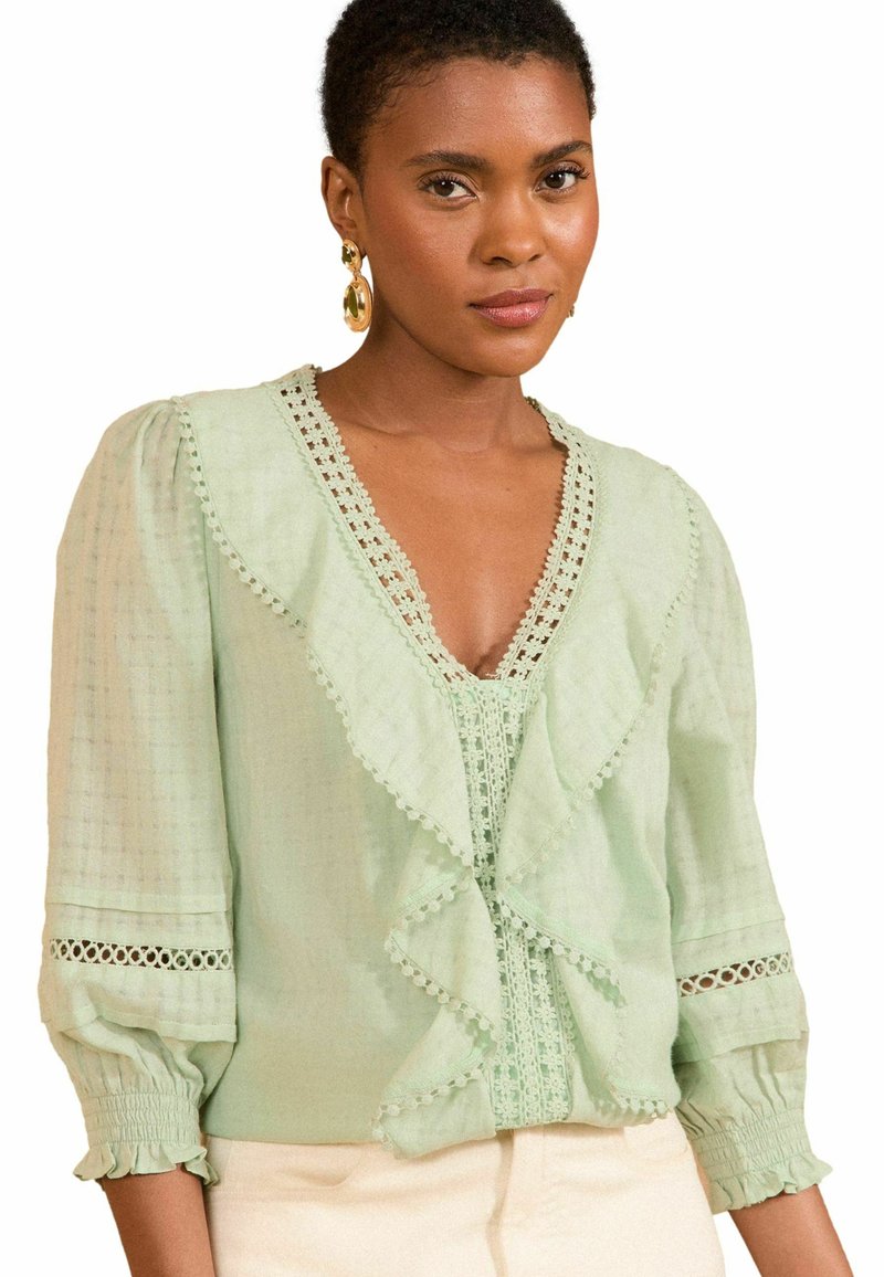 Blusa verde chiaro con scollatura a V, parte anteriore arricciata e delicata rifinitura in pizzo, maniche a sbuffo e polsini arricciati. Il tessuto appare leggero.