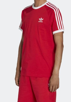 Homme portant un t-shirt rouge Adidas et un short avec bordure blanche et trois bandes blanches sur les manches, sur un fond uni.
