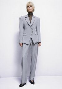Szürke, szabott öltöny, strukturált blézerrel és bőszárú nadrággal. A blézeren két gomb, szögletes zsebek és sima anyagú kivitel található.