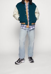 Giacca varsity in lana teal con maniche crema, camicia a quadri, top beige, jeans chiari e sneakers nere/metalliche. Sovrapposizioni per il freddo.