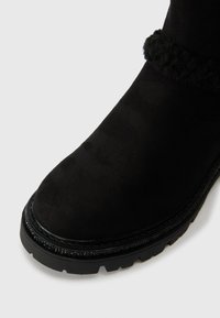 Fekete bokacsizma suede-szerű textúrával, puha, bolyhos gallérral, vastag fekete talppal és díszítő varrással a széleken.