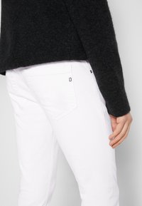 Jeans blancos con detalle en el bolsillo trasero, combinados con un suéter negro de textura. Enfoque en las texturas de la tela y el contraste de colores.