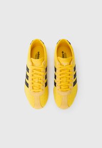 Κίτρινα σουέντ αθλητικά παπούτσια με τρεις μαύρες ρίγες, υφασμένο σχέδιο και κορδόνια που ταιριάζουν. Φέρουν το λογότυπο της Adidas στη γλώσσα και στο εσωτερικό της σόλας.