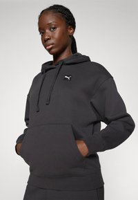 Schwarzer Hoodie aus weichem Material mit einer Kängurutasche, Kapuze mit Kordelzug und einem kleinen weißen Puma-Logo auf der Brust.