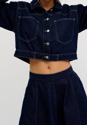 Femme portant une veste en jean courte bleu foncé avec des boutons et une jupe en jean assortie taille haute, montrant le ventre sur un fond uni.