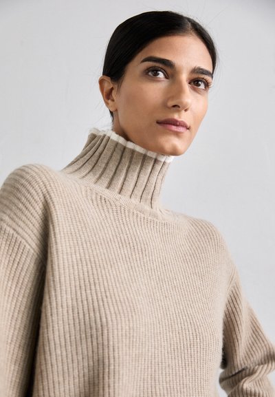 Beige gerippter Rollkragenpullover mit hohem Kragen und weißen Akzenten am Rand. Weiche Textur mit weiten, lockeren Ärmeln.