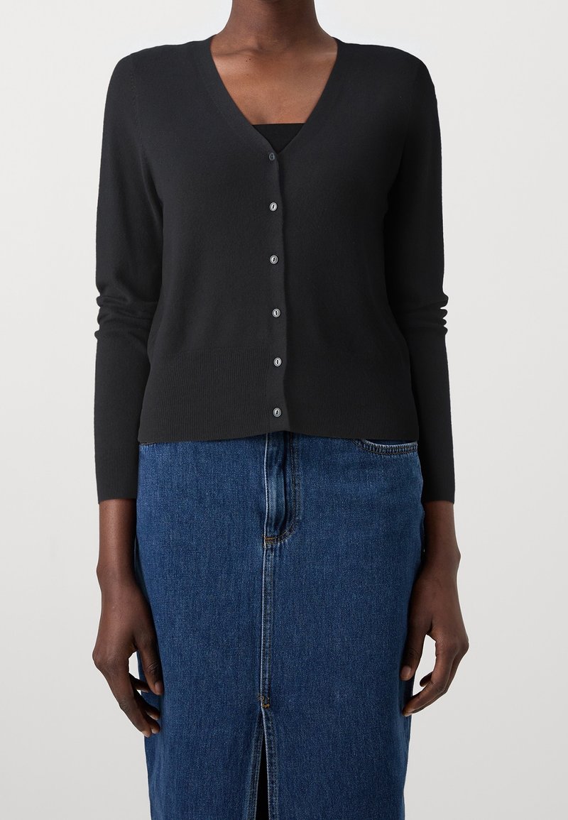 Cardigan noir à col en V boutonné porté sur un haut noir, associé à une jupe en jean bleue taille haute avec une fente à l'avant, portée par une personne debout.