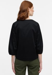 Blusa nera con una vestibilità ampia, scollo rotondo e maniche a tre quarti a sbuffo. Tessuto morbido con una leggera lucentezza. Abbinata a pantaloni verdi.
