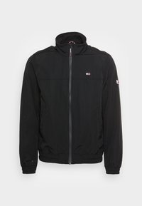 Svart lättviktig jacka med hög krage, hel dragkedja fram, elastiska muddar och en liten logotyp på bröstet. Strukturerad tyg.